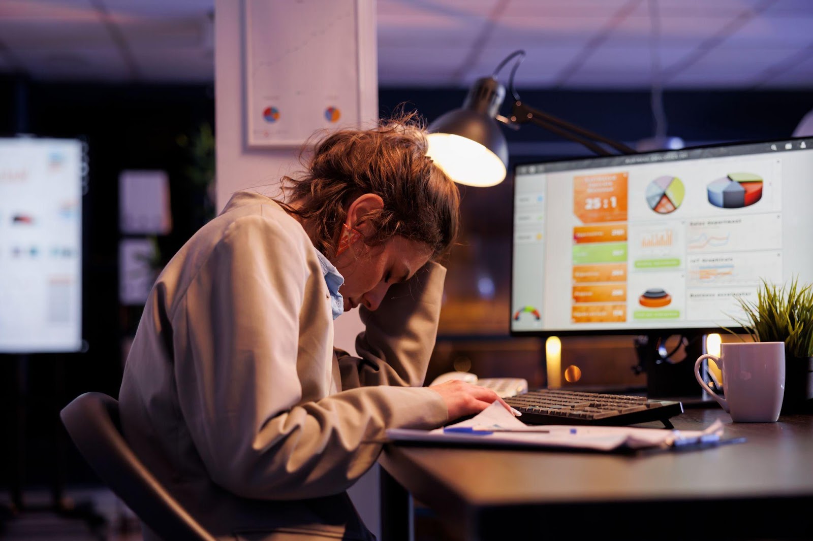 A tired office worker sitting at a desk late at night, leaning on their hand while surrounded by paperwork and a glowing computer screen. The image captures the exhaustion of working overtime.