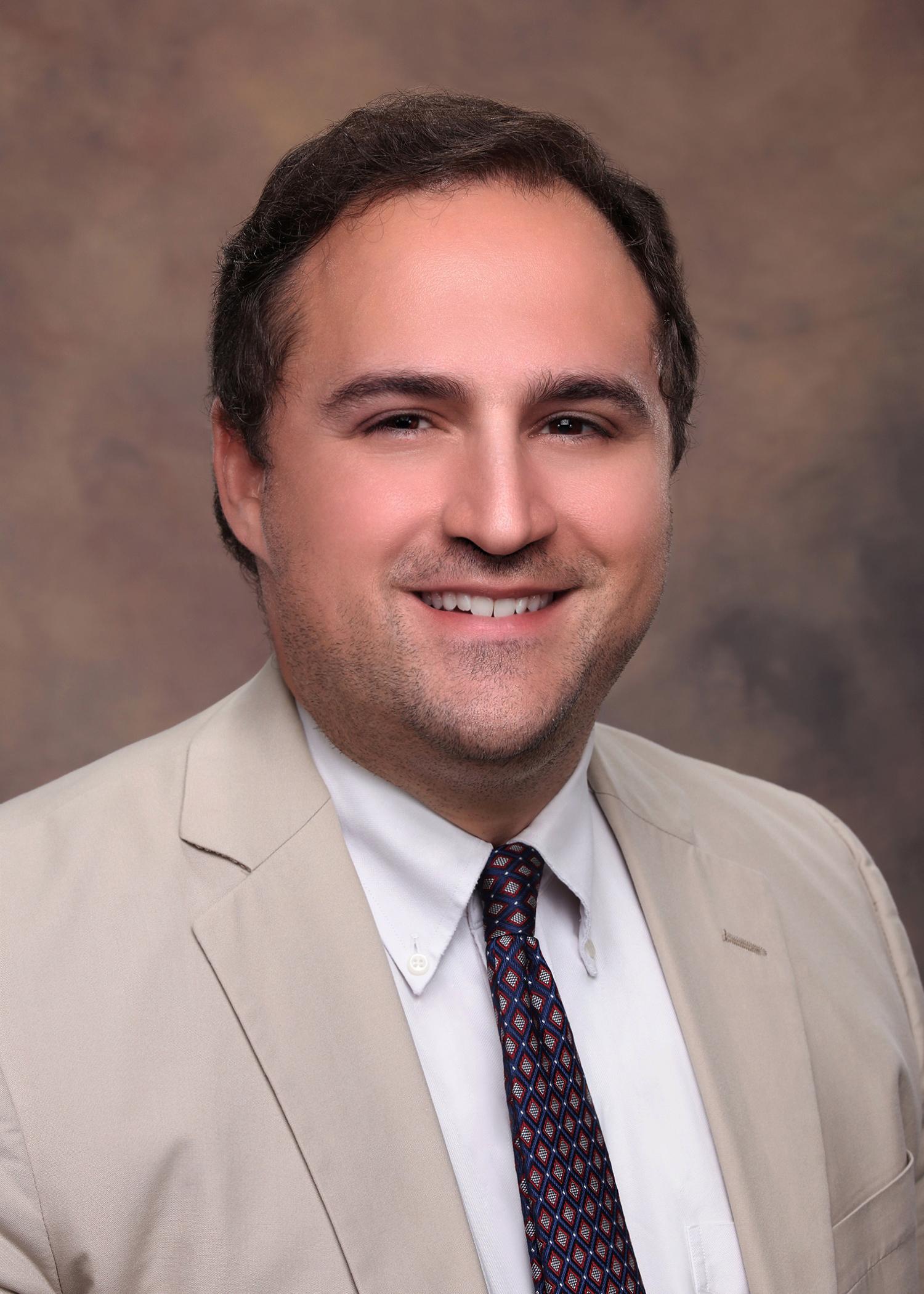 Adam Maniscalco, a divorce attorney practicing with Baxley Maniscalco, LLP, poses for a picutre in a beige blazer, a red tie, and a white shirt.