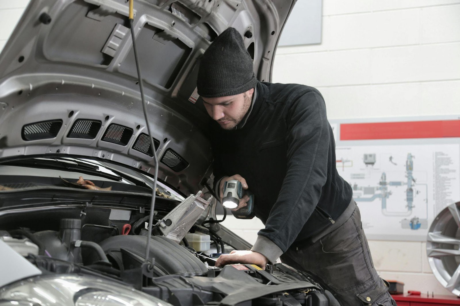 Mechanic inspecting car engine with open hood, holding diagnostic tool and flashlight.