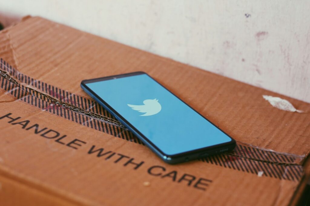 An iPhone sits on top of a cardboard box. The iPhone has the Twitter app opened, and the top of the box reads “Handle With Care,” symbolizing the attentiveness required to understand social media’s impact on personal injury claims.