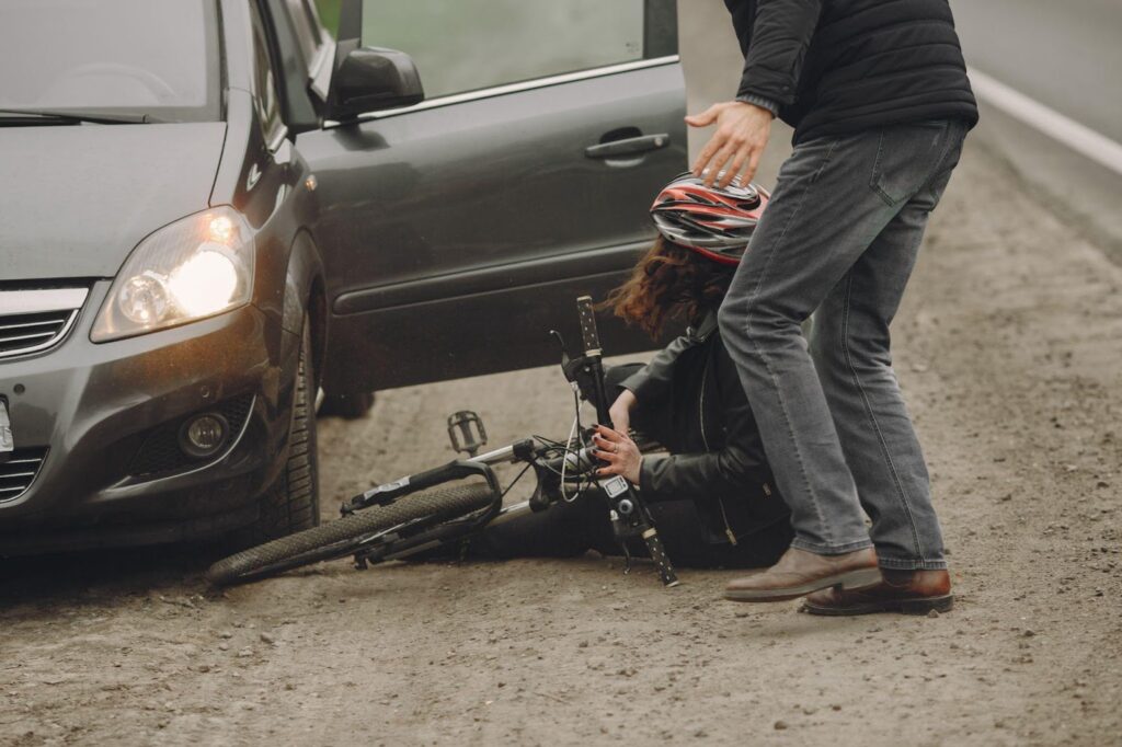 A woman cyclist lies on the road, visibly injured, with her bicycle. A car owner, who appears distressed and concerned, stands beside the cyclist. The scene depicts the aftermath of an accident involving the car and the cyclist.