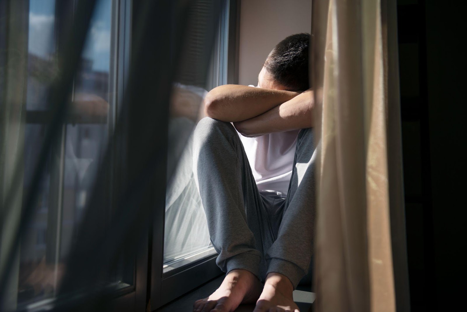 Person sitting on window sill, head buried in arms, conveying distress and isolation after traumatic experience.