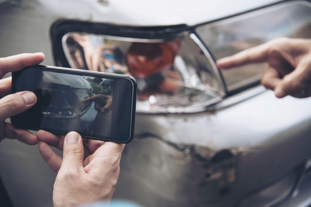 Hand holding smartphone, photographing crumpled rear bumper of car after a rear-end collision.