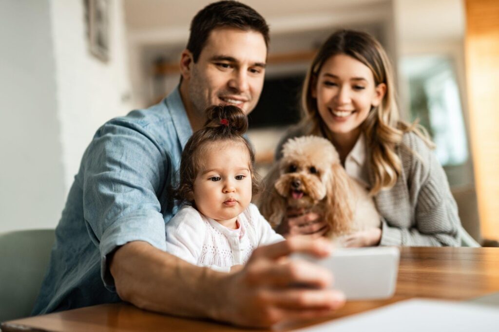 A family of two parents, one child, and a dog are smiling as they take a selfie together.