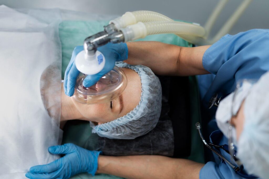 An anesthesiologist in a surgical gown and mask carefully administering anesthesia to a patient through a mask, while the patient lies on the operating table, preparing for surgery.