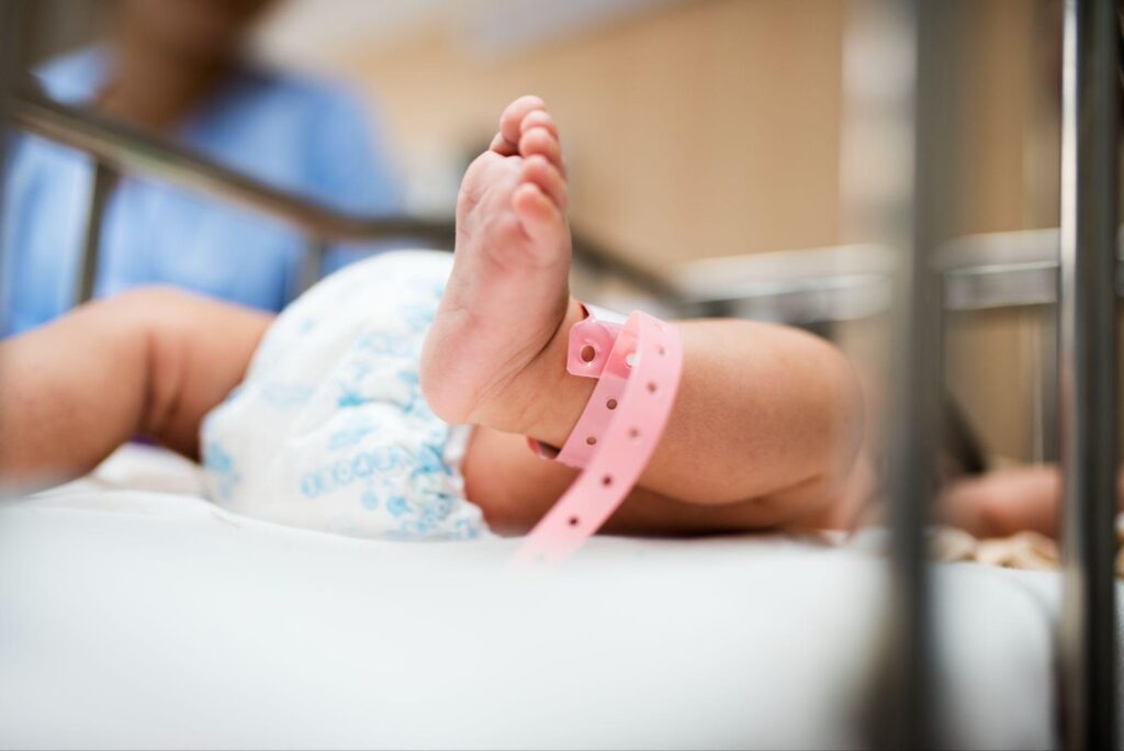 A newborn baby lying in a hospital bassinet with a pink identification bracelet around its ankle. The infant's legs are visible, and a healthcare professional is blurred in the background. The image relates to birth injuries, highlighting the vulnerability of newborns during the delivery process.