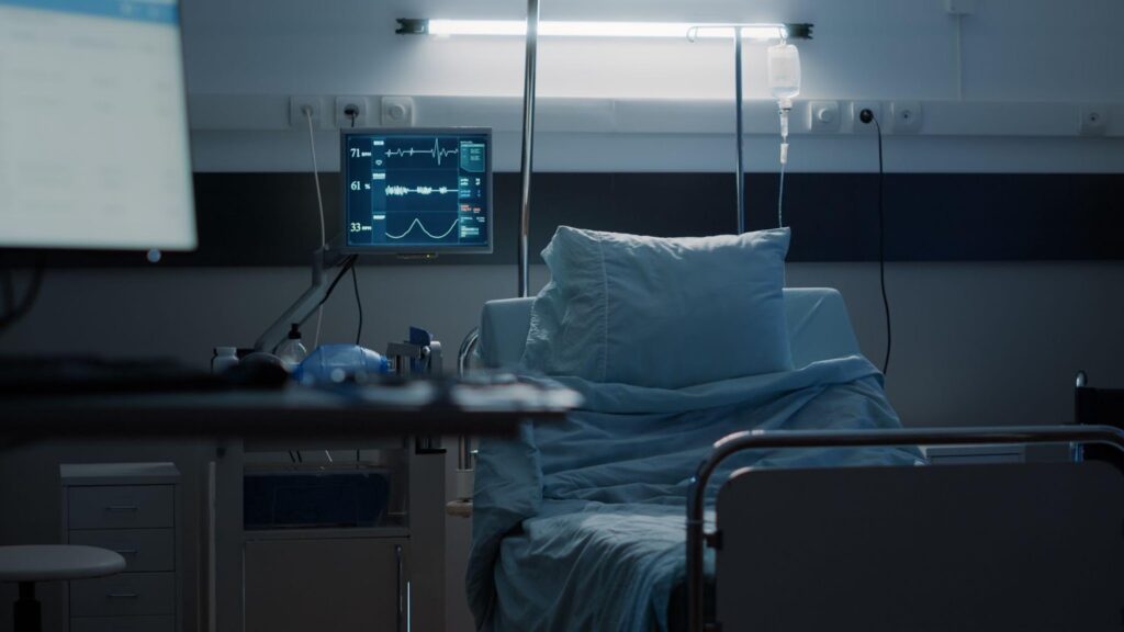 An empty hospital bed in a dimly lit room, with medical monitoring equipment displaying vital signs on a screen.