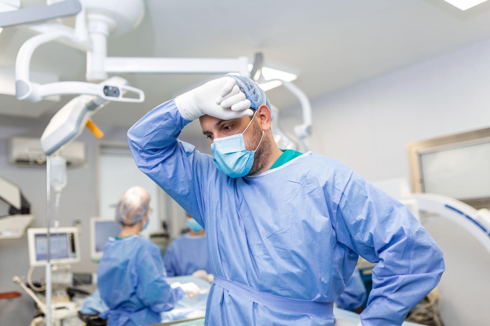 A distressed doctor stands in a surgery room, appearing deep in thought, with medical equipment in the background. The image conveys the high-stress environment of surgical procedures, underscoring the serious implications of surgical errors and the potential for medical malpractice.