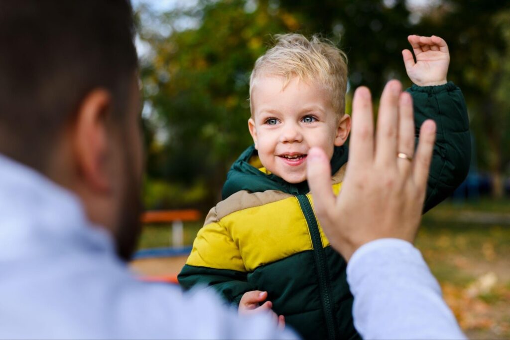 A smiling child raising his hand to high-five his dad, who adopted him, as they stand together outside. This joyful moment highlights the strong bond between them and reflects the journey of adoption, while also symbolizing the choice between open adoption and closed adoption.