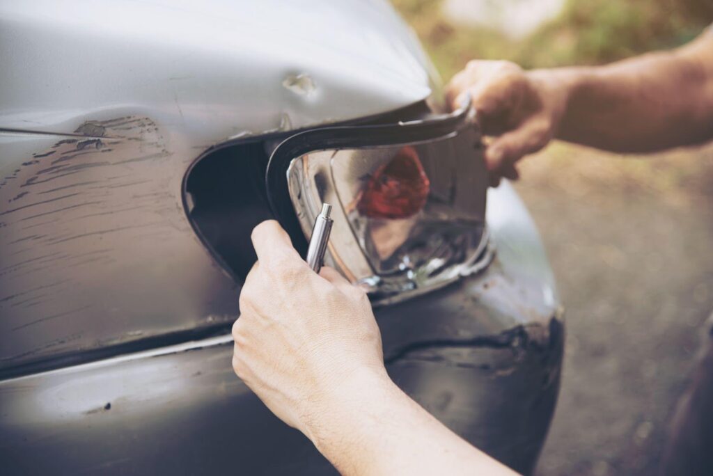 A hand carefully fixes the front light of a car that has been involved in a rear-end collision. The light appears damaged, with slight dents on the vehicle’s front, indicating the aftermath of the accident and the process of repairing the car.