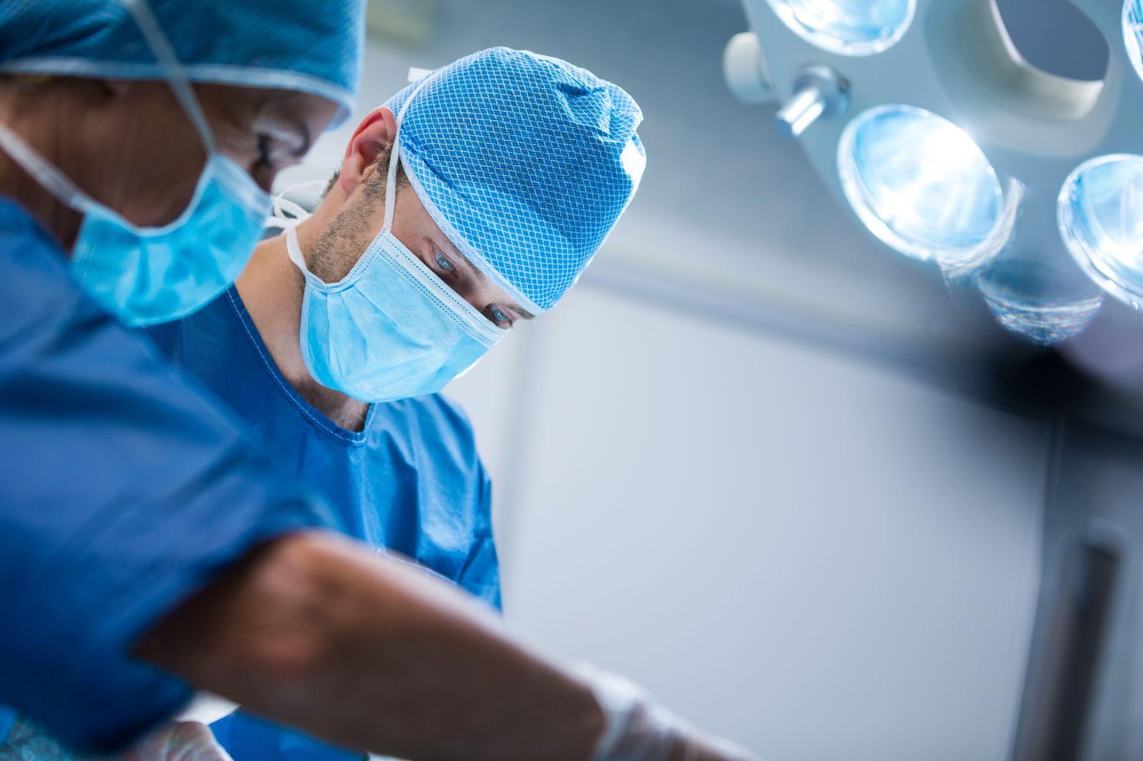 Two surgeons in surgical gowns and masks performing an operation under bright overhead lights, surrounded by medical equipment in a sterile operating room.