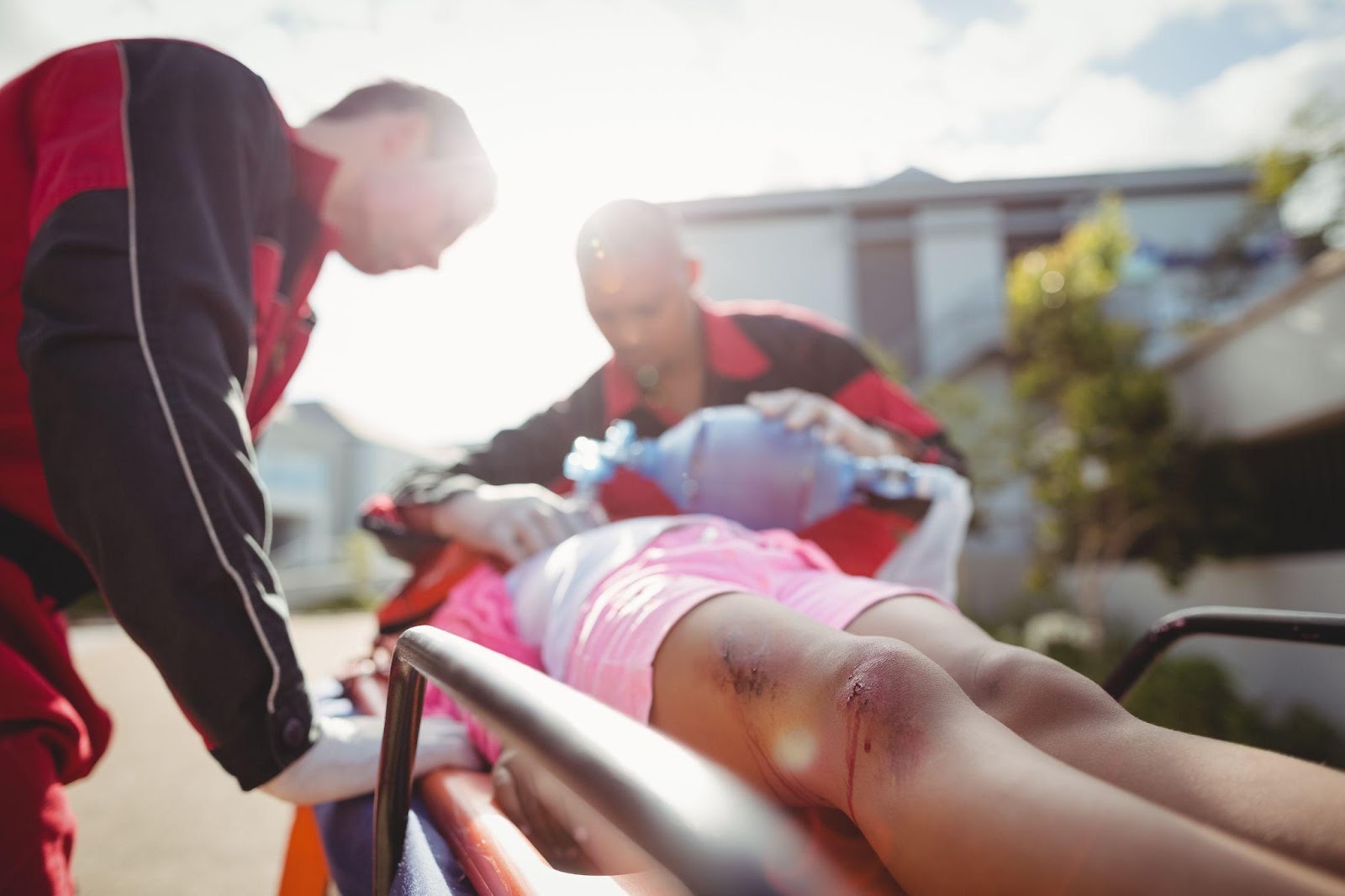 Paramedics attend to an injured girl on a stretcher, administering oxygen as she lies with visible scrapes and burn injuries on her knees.