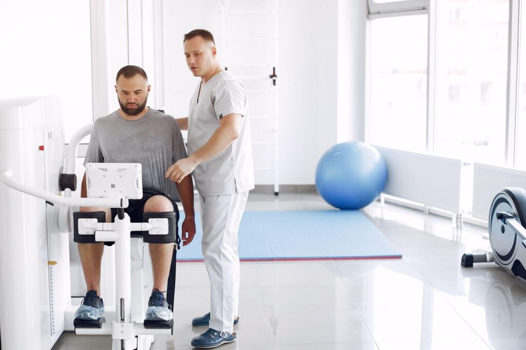 A physical therapist assists a male patient using an exercise machine in a rehabilitation clinic, demonstrating the use of adaptive devices for recovery.