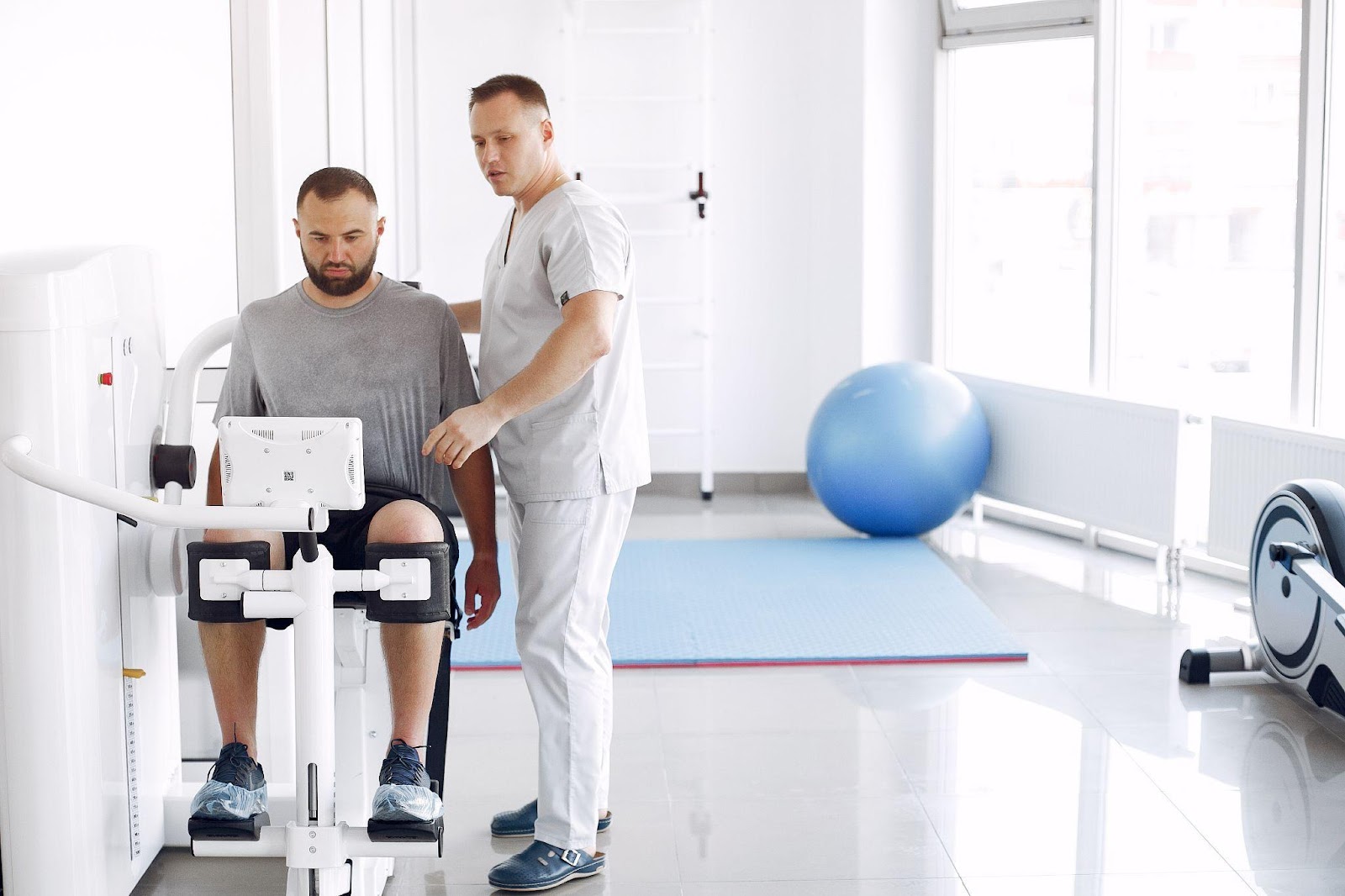 A physical therapist assists a male patient using an exercise machine in a rehabilitation clinic, demonstrating the use of adaptive devices for recovery.