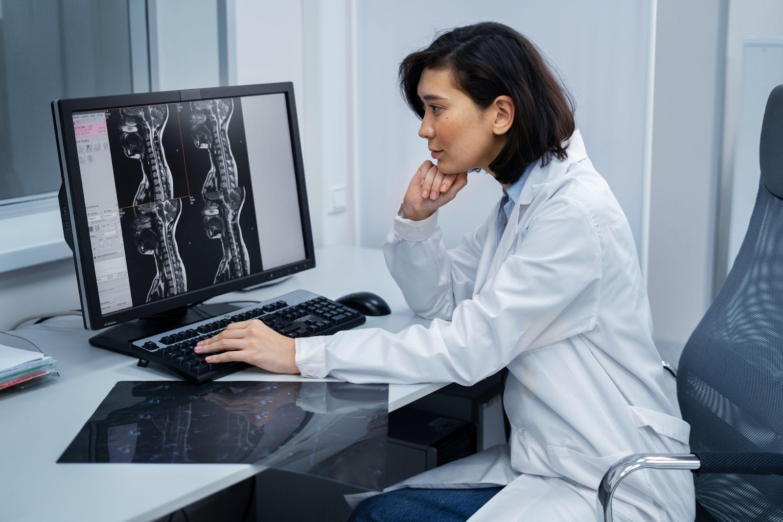 A doctor sits at her desk, studying a CT scan of a spinal cord displayed on her computer screen, focusing on detailed spinal analysis and diagnosis.