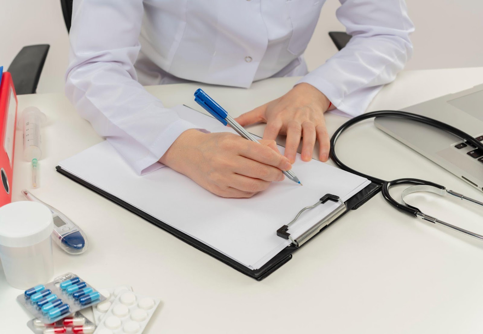 Close-up of a doctor's hand writing a prescription on paper, with a pen in hand. Pills are scattered on the desk, highlighting issues related to medication, prescriptions, and patient care.