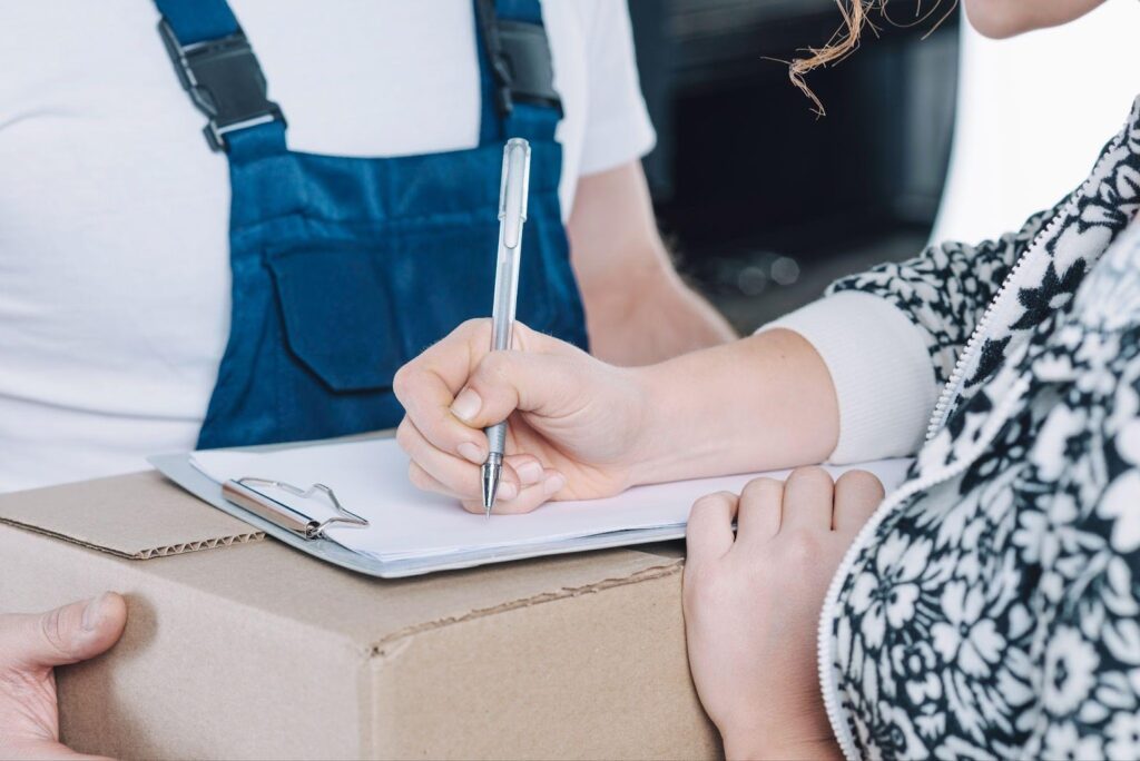 A woman signs documents related to product liability, with a box containing the product in front of her. The image represents the legal process involved in addressing issues of product responsibility and consumer safety.