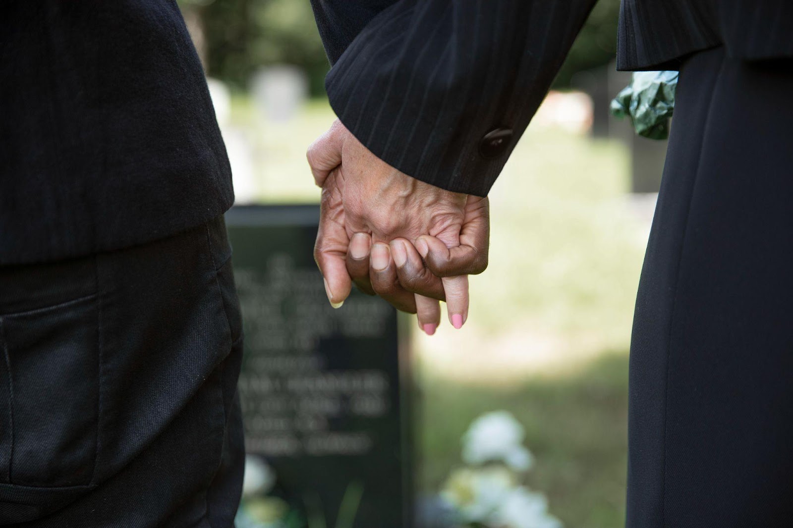 A close-up of a couple holding hands in front of a tombstone, with the stone visible in the softly blurred background. The image reflects shared grief and the bond between loved ones as they remember a life lost.