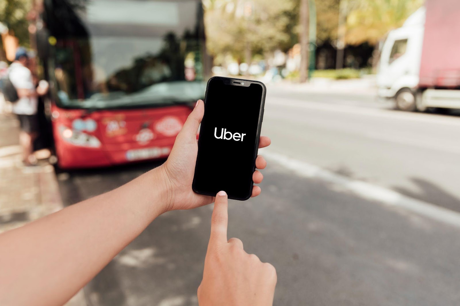 A person holding a smartphone with the Uber app open, standing near a city street with a bus and vehicles in the background. This image represents the context of rideshare driver liability, emphasizing the importance of understanding legal responsibilities and protections when using services like Uber, Lyft, and similar platforms.