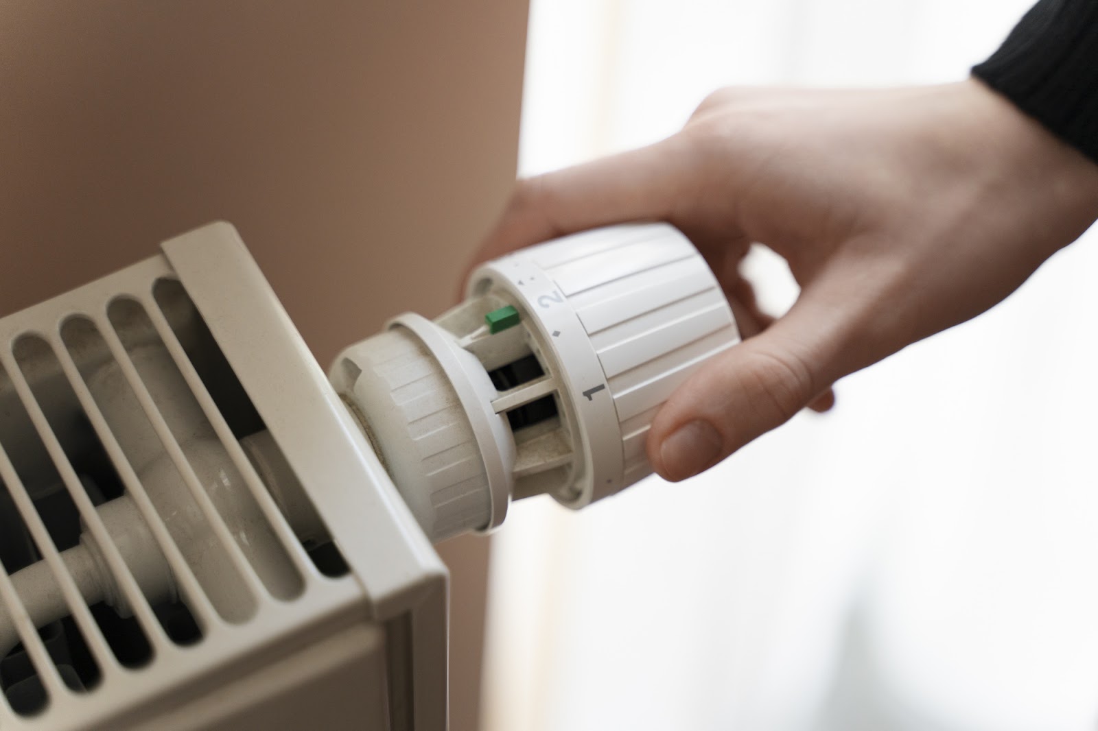 Close-up of a hand adjusting a white thermostatic valve on a home radiator with vertical slots. This heating equipment requires proper maintenance and carbon monoxide detectors to prevent CO poisoning, ensure safety compliance, and protect occupants' legal rights.