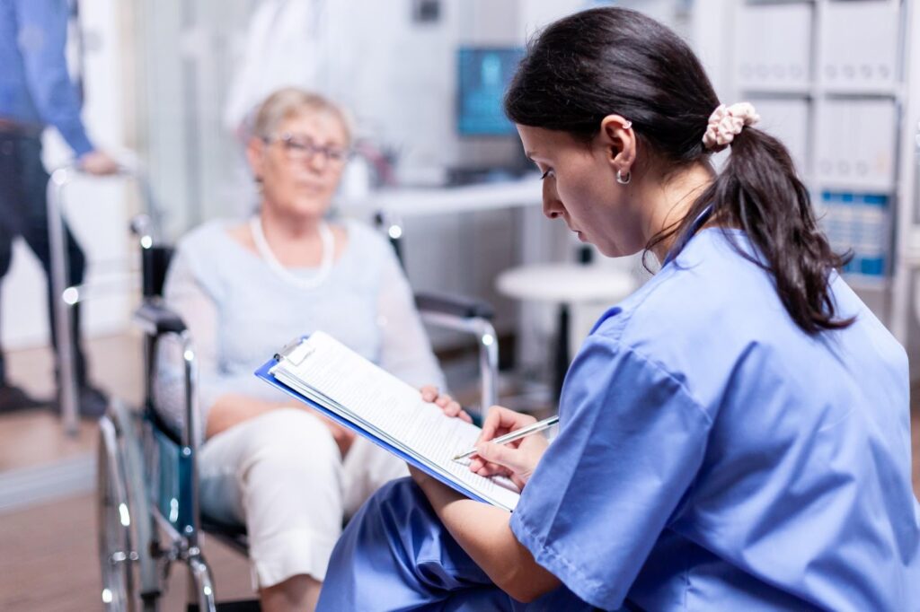 An image depicting a healthcare professional attentively taking notes while seated near an elderly woman in a wheelchair. This highlights the importance of vigilance in identifying signs of elder abuse in nursing homes, ensuring proper reporting, and taking legal action to protect vulnerable individuals.