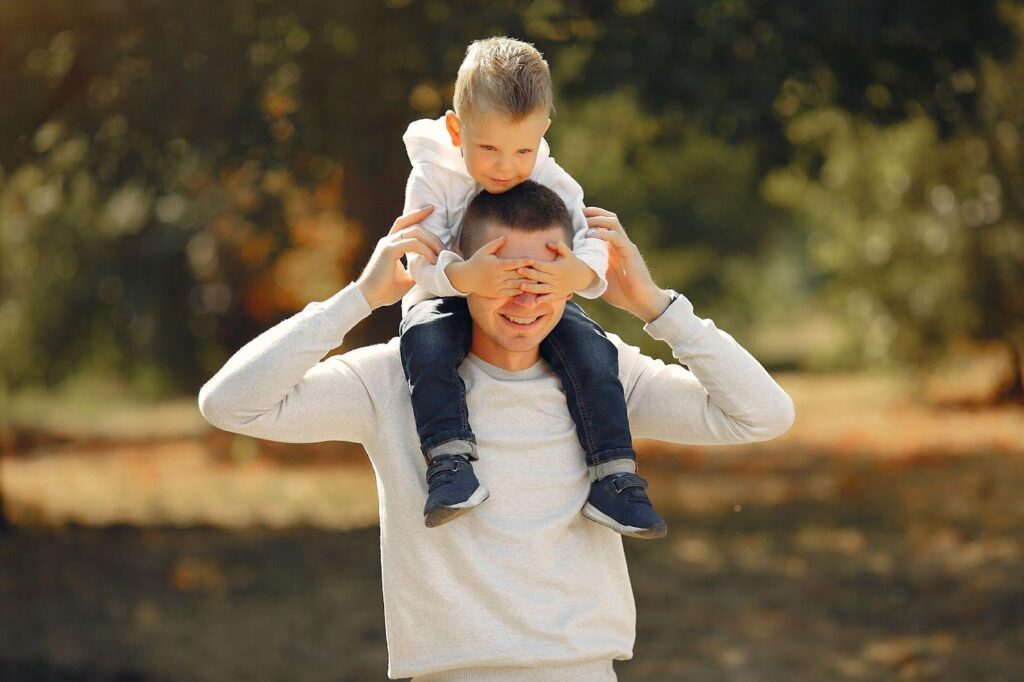 A father carrying his child on his shoulders, both smiling and enjoying an outdoor moment. The image conveys the topic of FAQs about paternity in Alabama, focusing on fatherhood and parental rights.