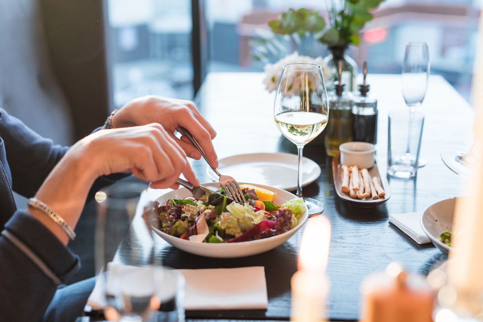 A person eating at an upscale restaurant, using utensils to cut into a colorful salad served in a white bowl. There's a glass of white wine, condiment bottles, and breadsticks on the dark wooden table. The scene emphasizes fine dining and careful food handling in a well-lit restaurant setting with windows in the background. The image relates to restaurant service and food safety considerations.