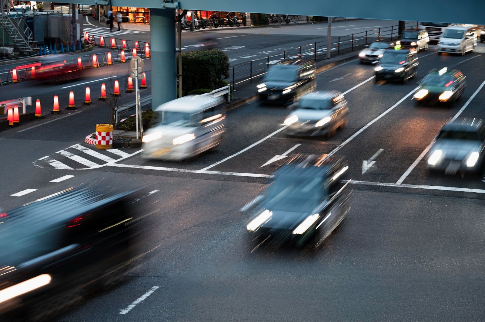 An aerial view of a busy intersection at dusk or night, showing cars in motion with blurred headlights and taillights indicating fast movement. The road is marked with traffic lanes and features orange safety cones and road barriers. The motion blur of vehicles suggests rapid traffic flow, while safety measures like road markings and barriers are clearly visible, highlighting the context of traffic safety and potential road incidents. The scene captures the dynamic and potentially dangerous nature of urban traffic conditions.