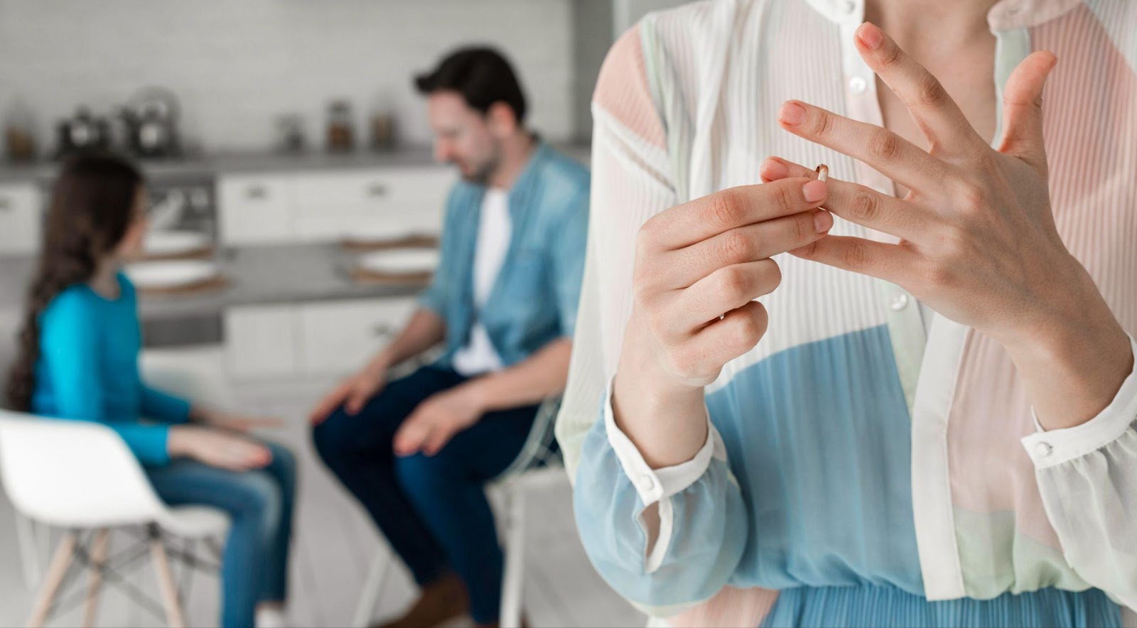 A woman removing her wedding ring in the foreground, with a blurred family scene in the background, symbolizing the complexities of separation and emphasizing the importance of hiring a lawyer for a high-asset divorce in Alabama.