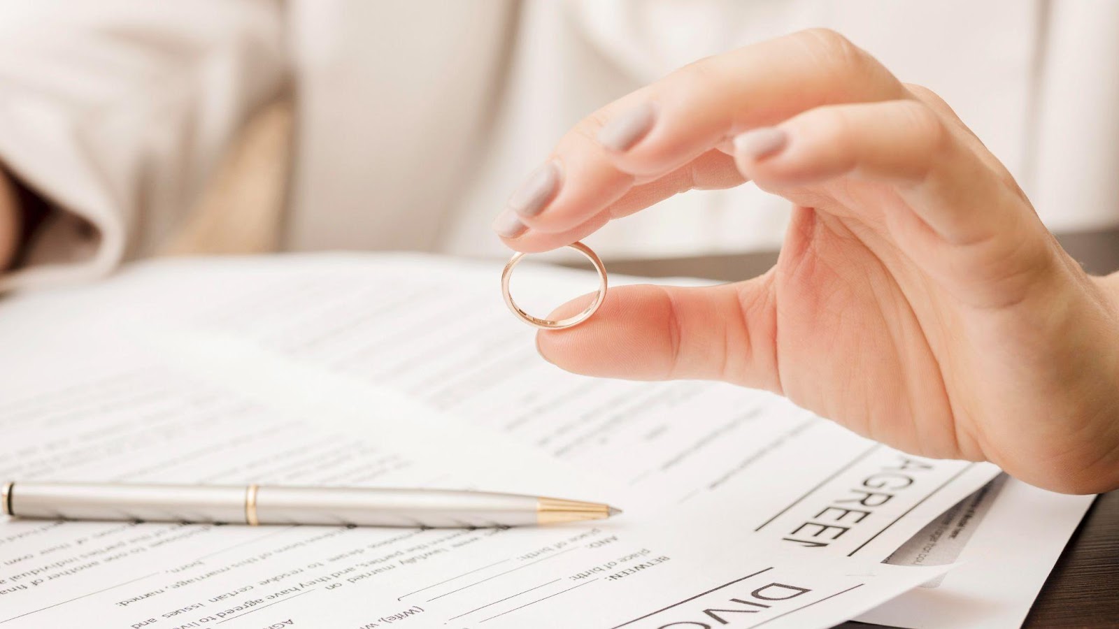 A hand holds a wedding ring over divorce paperwork, with a pen in the background.