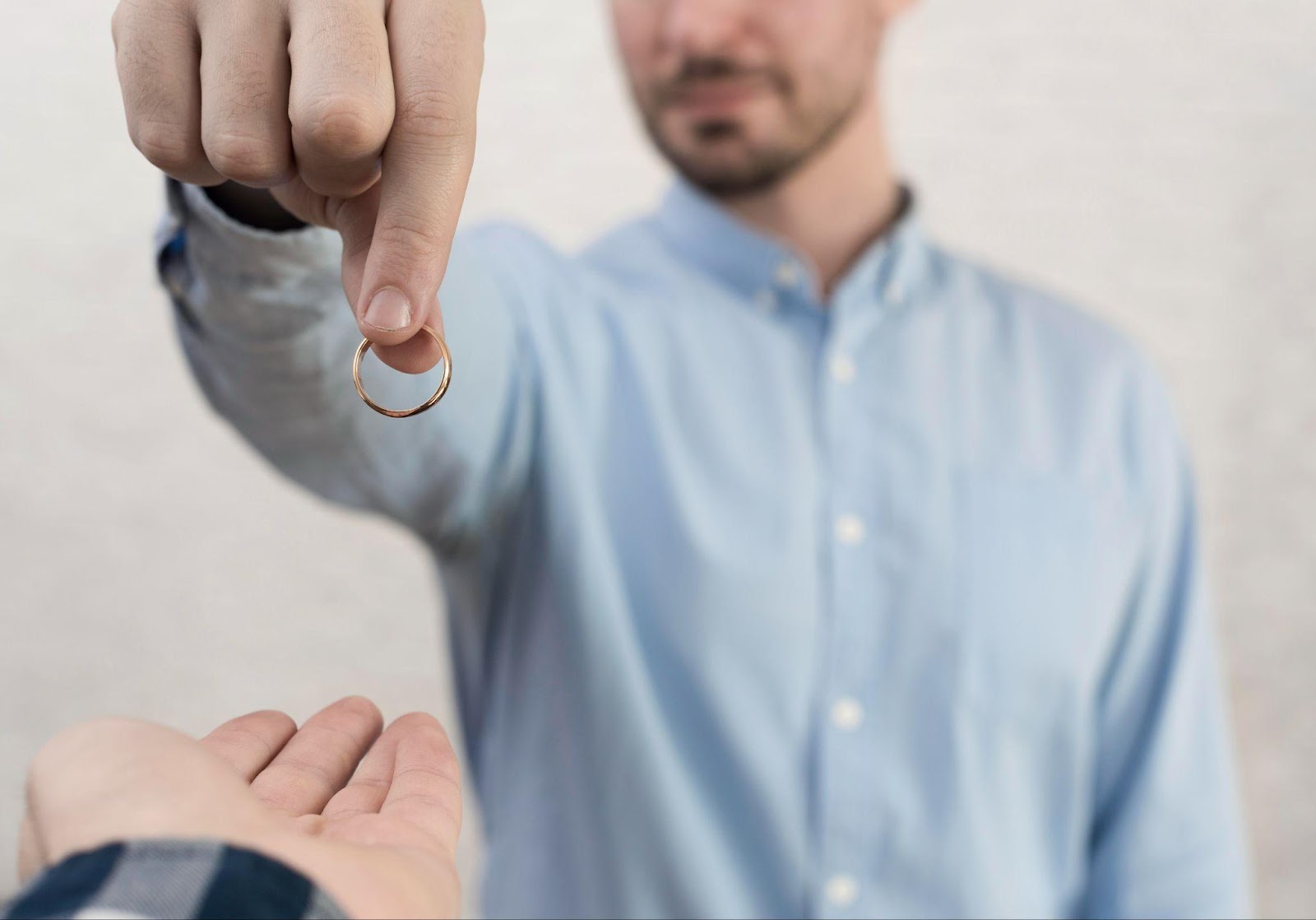 Close-up of a man in a light blue shirt holding out a wedding ring, symbolizing divorce, separation, or the end of a relationship.