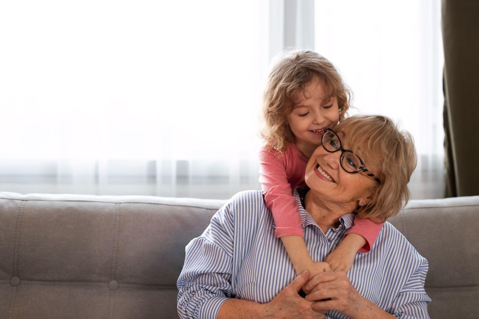 A smiling grandmother in a blue and white striped shirt and glasses embraces her young granddaughter, who is wearing a pink shirt. They are sitting together on a gray couch, sharing a warm, affectionate moment with the child hugging her grandmother from behind.