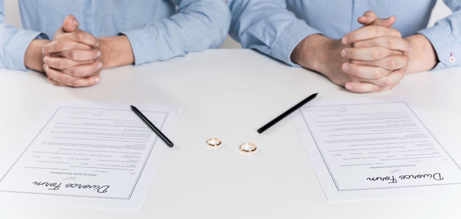 Two people sitting at a table with divorce papers, wedding rings, and pens, symbolizing the process of ending a marriage.