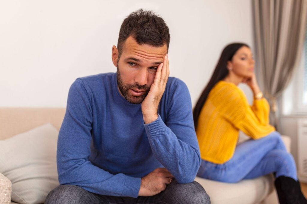 A distressed man sits on a couch, his face resting in his hand, while his partner turns away, highlighting the emotional toll of infidelity in marriage. When adultery becomes a factor in divorce, it can impact property division, alimony, and child custody.