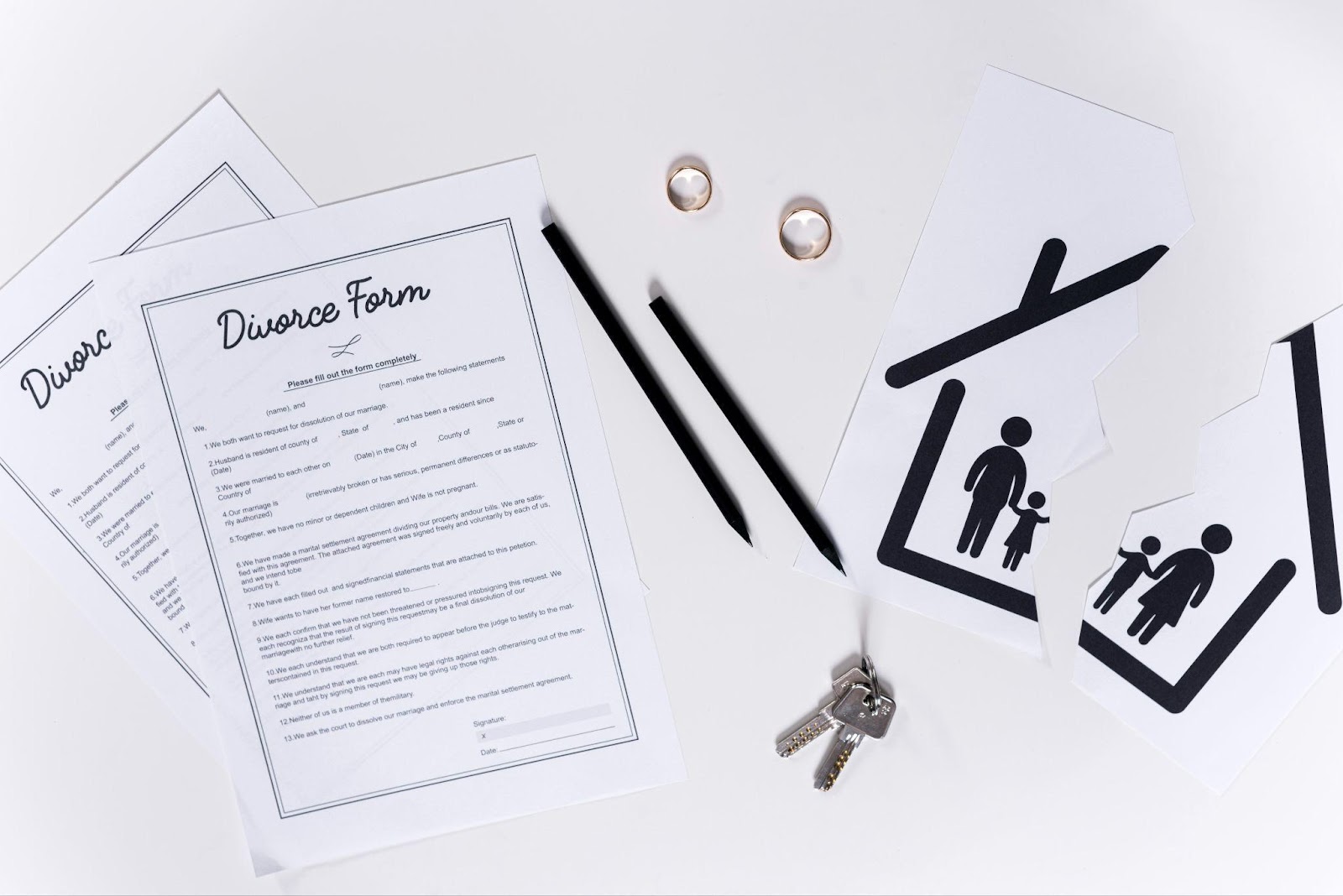 Flat lay of divorce paperwork including divorce forms, two wedding rings, house keys, and family pictogram symbols suggesting separation or division of family unit. The image includes black pens and is set against a white background.