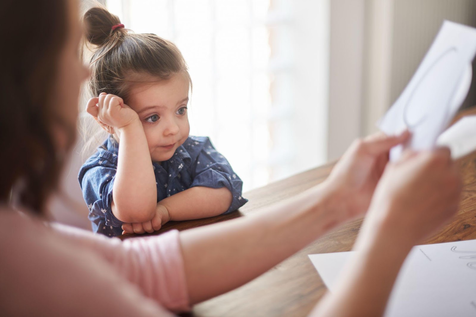 A young girl with her hair in a top knot sits at a table, resting her head on her hand and looking thoughtfully to the side. An adult's hands can be seen holding a document in the foreground, suggesting a serious or contemplative moment between a child and an adult.