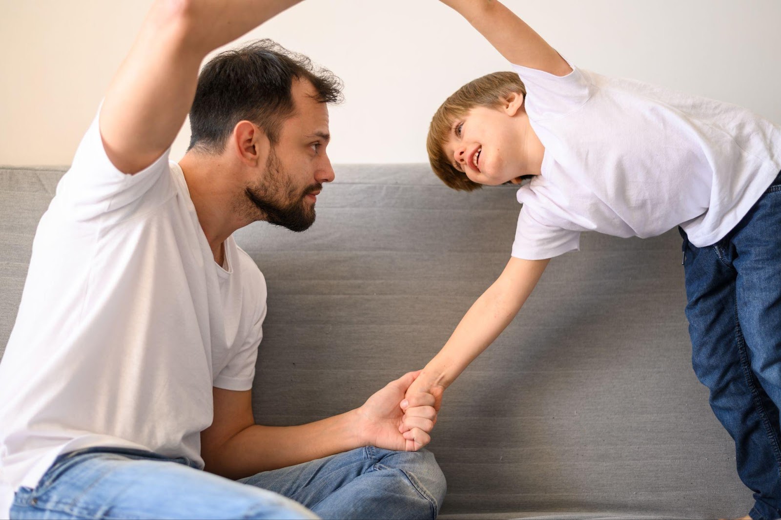 A father and son engage in a playful moment, with the father sitting on a gray couch and holding his son's hand. The son leans towards his father with a smile, while both have their other hands raised, suggesting a lighthearted interaction between parent and child.