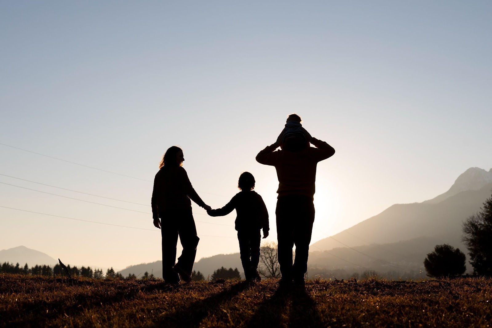 Silhouette of a family at sunset - a parent holding hands with a child in the middle while another parent carries a child on their shoulders, standing on a grassy hill with mountains in the background.