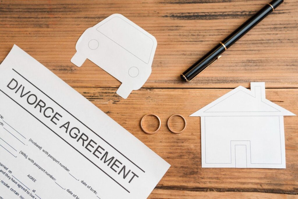 A divorce agreement document on a wooden table with paper cutouts of a house and car, two wedding rings, and a black pen, representing property division during divorce proceedings.