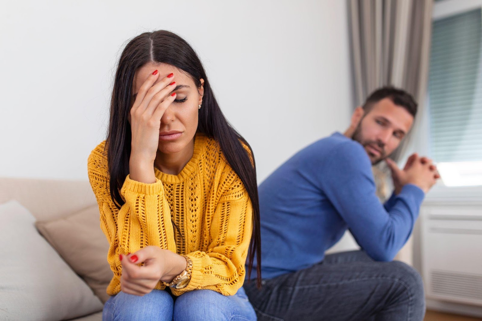 A distressed couple sitting apart on a couch, showing signs of relationship strain. A woman in a yellow sweater covers her face with her hand while wearing red nail polish, and a man in a blue sweater sits turned away in the background, both displaying body language suggesting emotional conflict or marital discord.