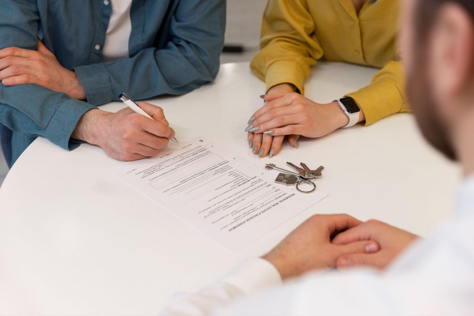 Two people sit at a white table, with one person in a blue shirt signing a document while another in a yellow shirt watches. A set of keys is placed on the document, suggesting a legal or contractual meeting, related to real estate.