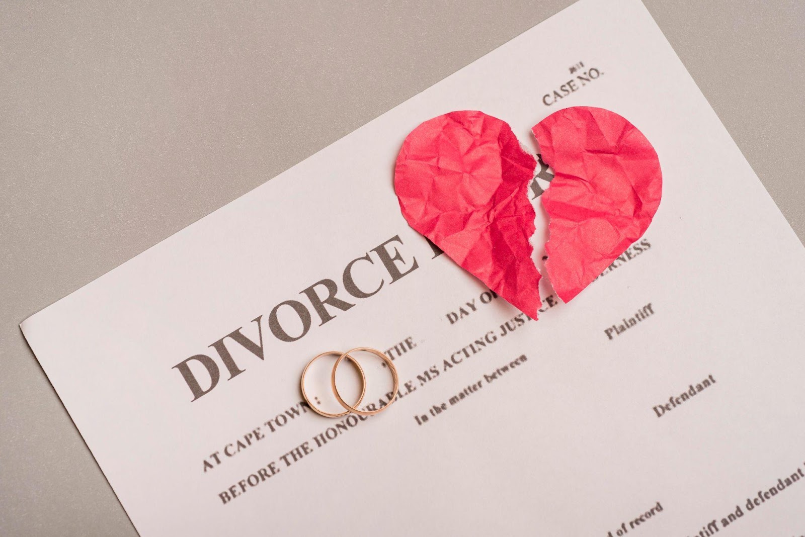 A divorce document with two wedding rings placed on it and a torn red paper heart above. The document shows "DIVORCE" in large text at the top, with legal text visible below.