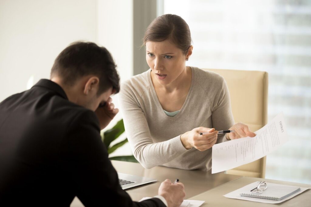 A woman in a beige sweater looking sternly at divorce paperwork while pointing at documents, sitting across from a visibly distressed man in a black suit during what appears to be a divorce consultation. A laptop and legal documents are spread across the desk between them.