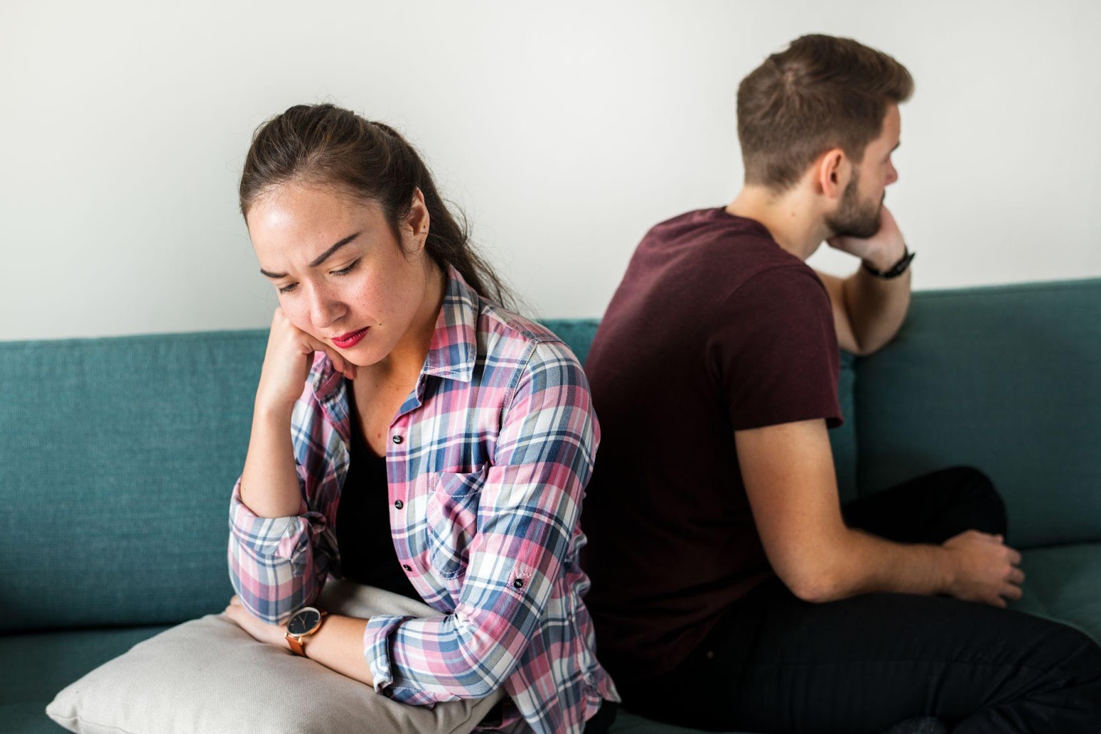 A couple sitting on opposite ends of a teal couch showing signs of relationship distress. A woman in a pink plaid shirt sits on the left looking downward with her hand on her face, appearing sad or worried. A man in a burgundy t-shirt sits on the right with his back partially turned, also with hand on face, suggesting tension or disagreement between them.