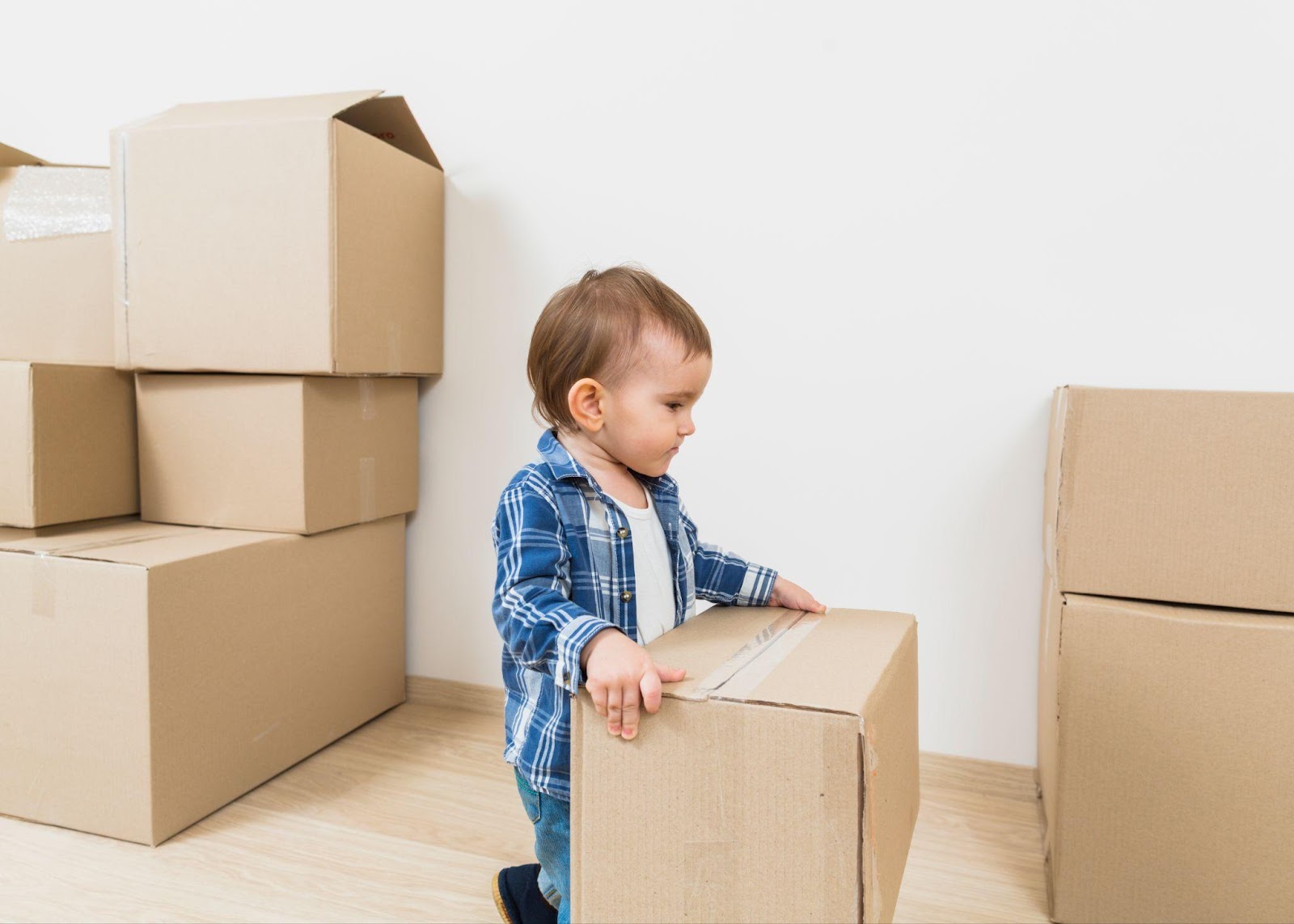 The image shows a toddler in a blue plaid shirt standing by moving boxes in an empty room with white walls. They're leaning against one of several cardboard boxes stacked around them.