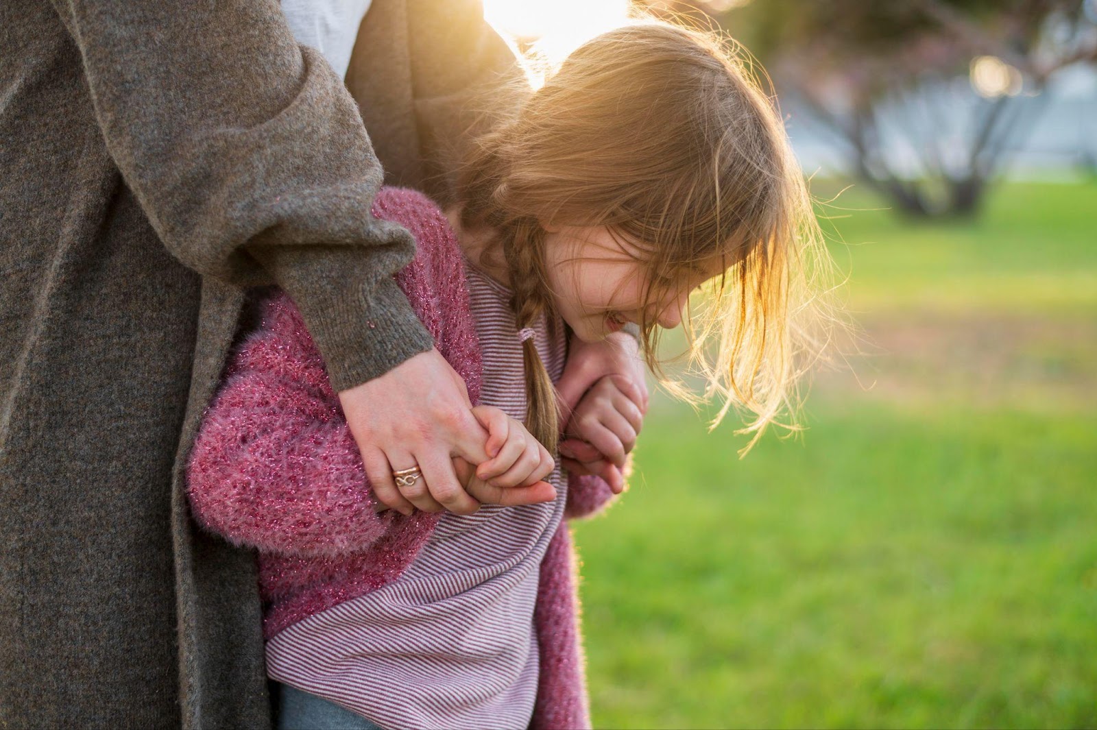 A young child with light blonde hair wearing a pink knit sweater and striped shirt stands close to an adult in a gray coat. The child is holding the adult's hand, which has a ring on it. They are outdoors in a park or grassy area with soft sunlight creating a warm backlit glow around the child's hair.