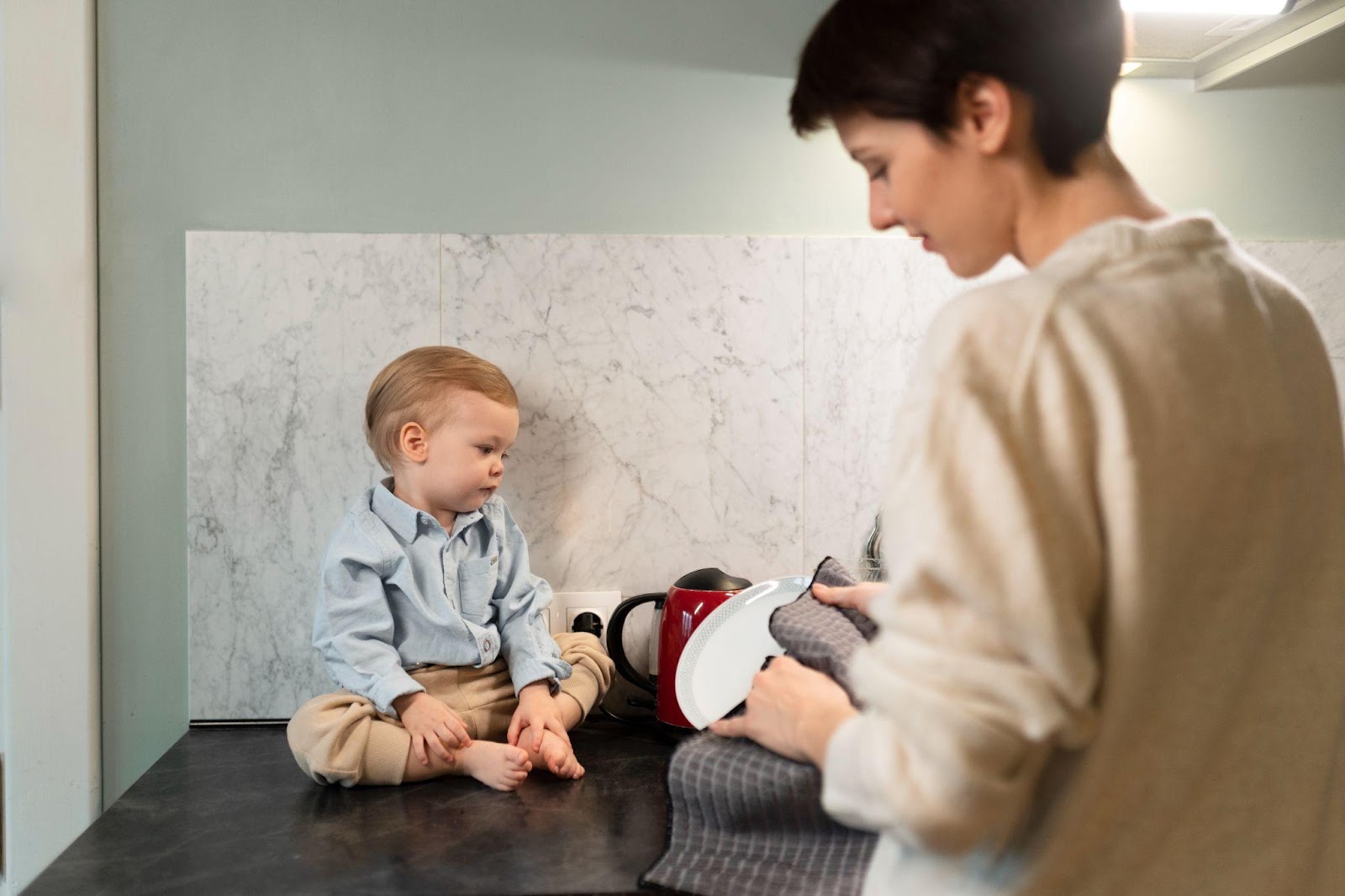 A young toddler with light brown hair wearing a blue button-up shirt and beige pants sits on a dark countertop against a marble wall. A person with short dark hair wearing a beige sweater stands nearby holding what appears to be kitchen items, looking at the child.