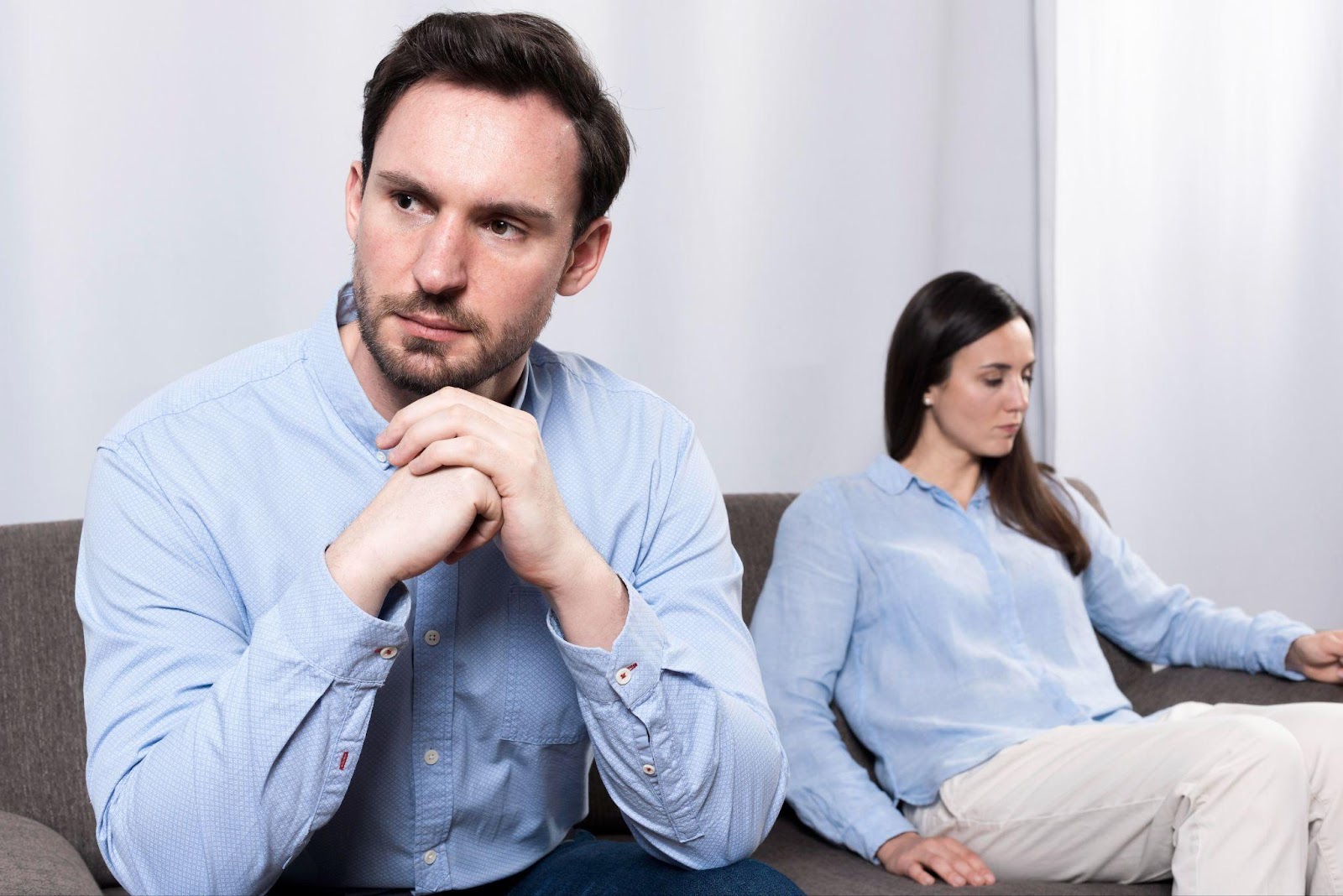 A man with short dark hair and a beard wearing a light blue button-up shirt sits pensively with his hands clasped, looking concerned. In the background, a woman with long dark hair wearing a similar blue shirt sits on the same couch but facing away from him.