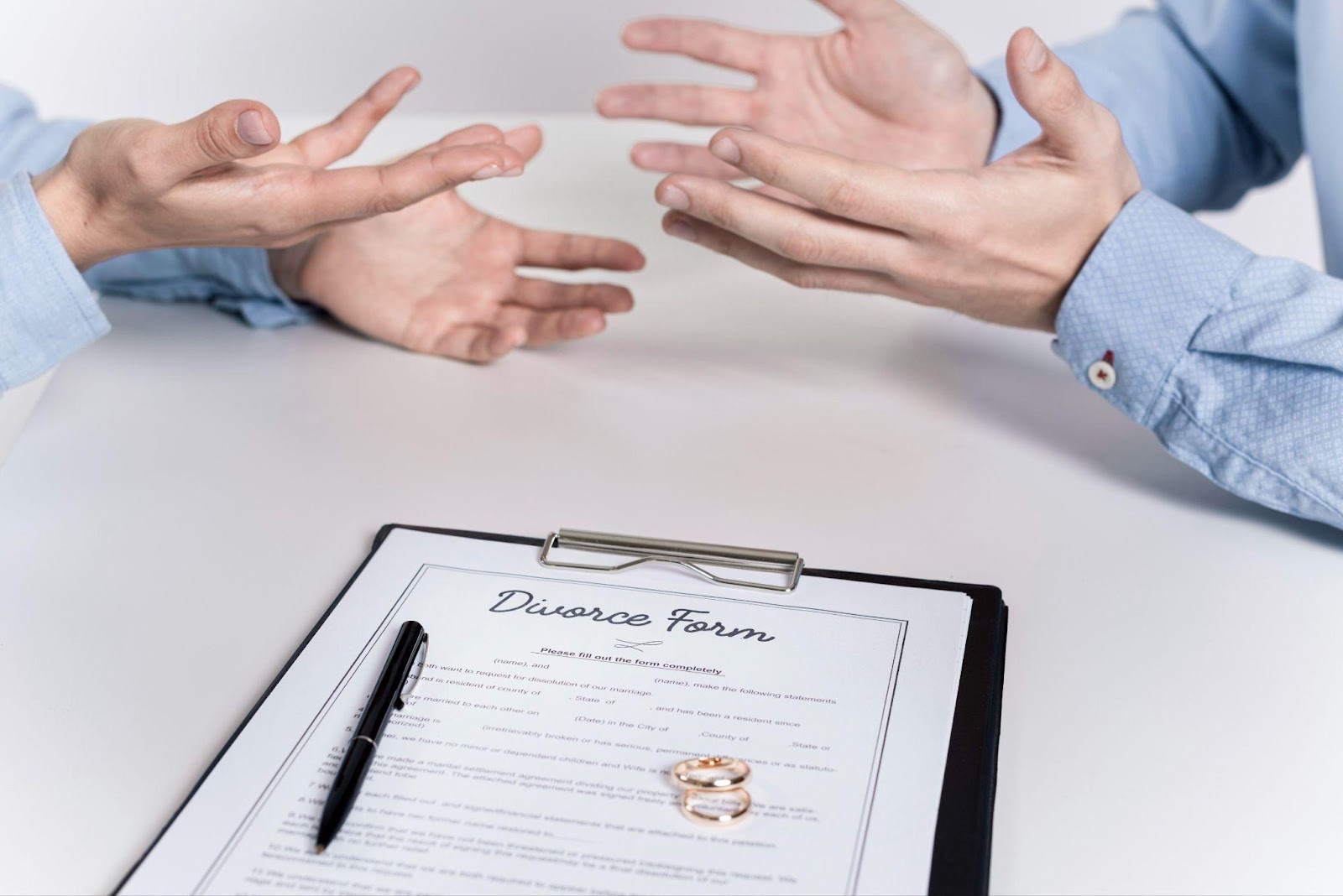 The image shows two people in light blue shirts gesturing with their hands during what appears to be a discussion about Alabama divorce, with a clipboard containing divorce paperwork, a pen, and wedding rings visible in the foreground.