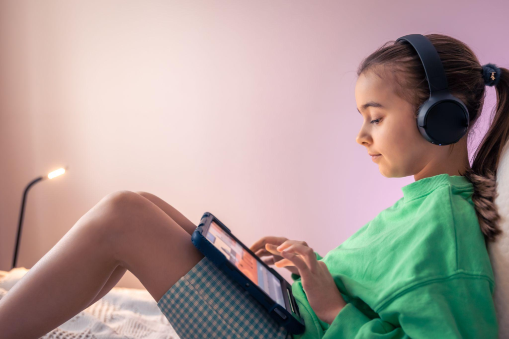 A girl wearing headphones and a green sweater uses a tablet while sitting on a bed.